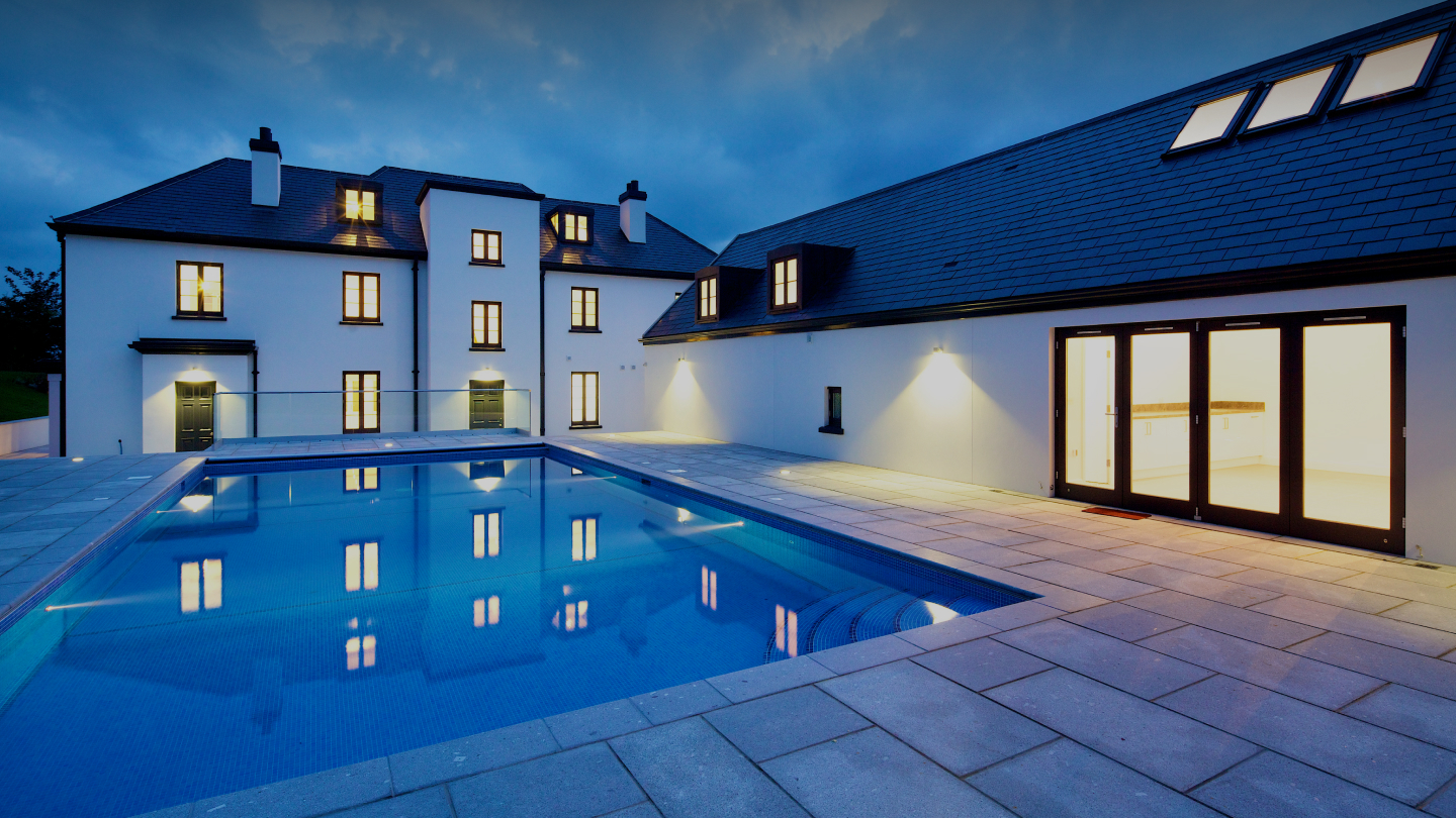 Large modern home with lit windows, slate roof and outdoor swimming pool at dusk